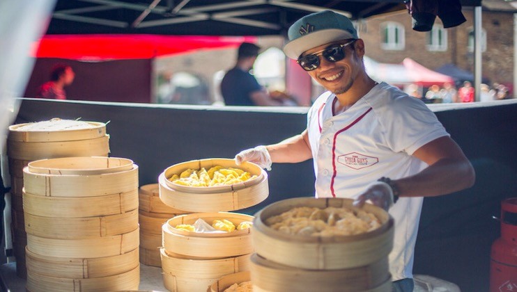 A smiling vendor wearing a cap and sunglasses holds a bamboo steamer filled with dumplings at a street market stall. Several other bamboo steamers filled with dumplings are stacked on the counter in front of him. The background shows other market stalls and people.