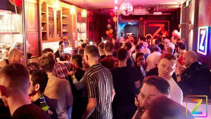 A crowded bar filled with people socializing and mingling under warm lighting. The bar area is busy with patrons, and the walls are adorned with colorful decorations, including balloons and a disco ball. A large Z is illuminated on the back wall.