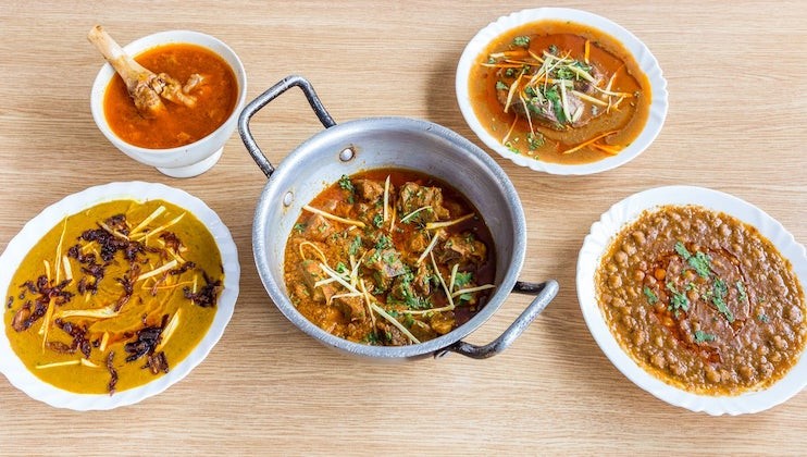 A spread of five dishes on a wooden table. The center features a metal pot with a meat curry garnished with julienned ginger and cilantro. Surrounding it are bowls of lentils, lamb curry, dal, and a rich, creamy mustard-based curry, all garnished similarly.