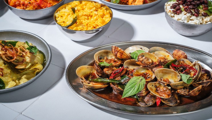 A plated dish of clams in a spicy sauce, garnished with basil leaves, is surrounded by bowls of vibrant foods including yellow rice, a vegetable and rice mixture, and a savory stew with vegetables, all set on a white tiled surface.