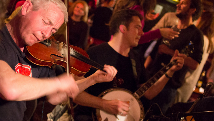 Two musicians passionately perform in a lively pub setting. The older musician on the left plays the violin with his eyes closed, while the younger musician on the right plays the banjo. The audience in the background appears engaged and joyful.