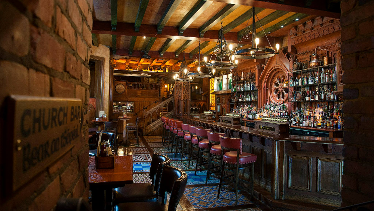 A cozy pub interior with warm lighting, wooden beams, and a long bar counter. The bar is lined with red cushioned bar stools and numerous bottles of liquor are displayed on shelves. A sign reading Church Bar is mounted on the brick wall near the entrance.