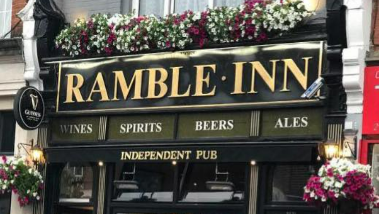 A pub with a black and gold sign that reads Ramble Inn and Independent Pub. The sign also advertises wines, spirits, beers, and ales. Flower boxes with pink and white flowers decorate the top and sides of the facade.