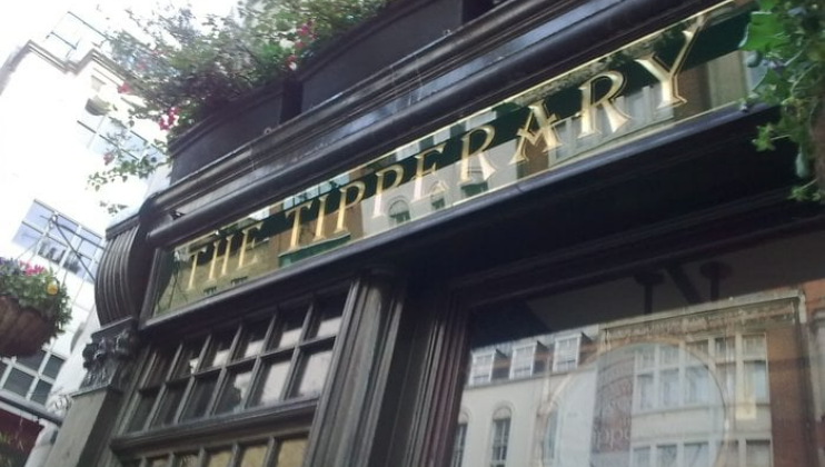 A photo looking up at the exterior sign of The Tipperary pub. The sign is gold with green background, and the building facade features ornate, dark wood framing with large windows. Flower boxes with greenery and pink flowers are partially visible at the top.