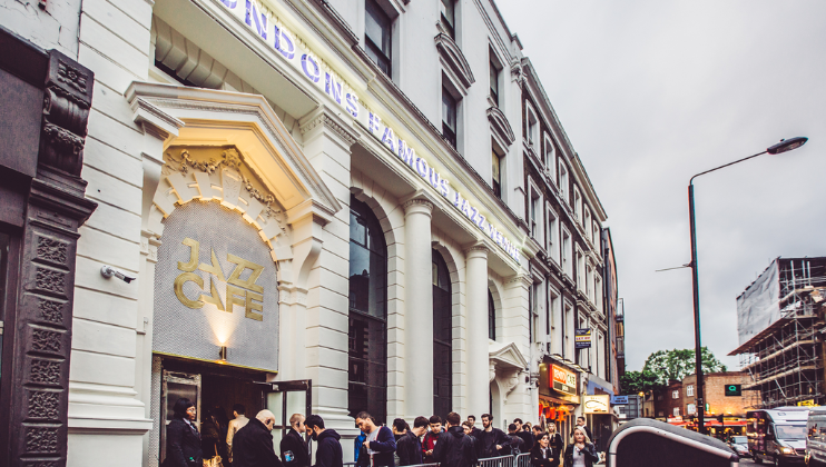 A group of people stand in line outside the entrance of a white, ornate building with LONDON'S FAMOUS JAZZ CAFE illuminated in large letters along the top. The surroundings include other buildings, street lights, and signs.