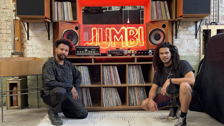 Two individuals are kneeling in front of a shelf filled with vinyl records. Behind them, a large neon sign with the word JUMBI is illuminated in red. The setup is an indoor space with a rustic brick wall, speakers, and audio equipment positioned above the shelf.