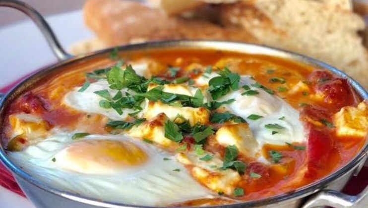 A shallow metal pan filled with a dish of baked eggs in a rich tomato sauce, garnished with chopped parsley. The dish is placed on a red napkin, and slices of bread are visible in the background.