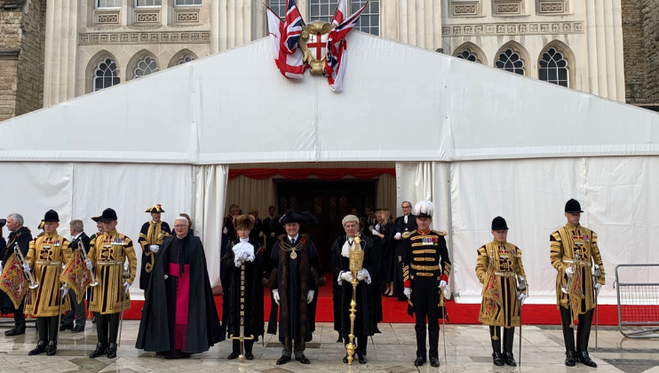 A group of people in ceremonial attire stands on a red carpet in front of a white marquee with British flags above it. They are flanked by individuals in uniforms carrying trumpets. The setting appears formal, possibly for a significant event or ceremony.