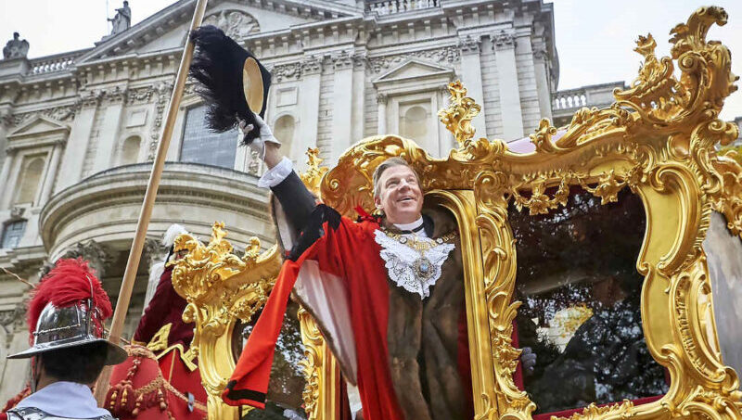 A person dressed in formal ceremonial attire and a red robe waves a black feathered hat from a lavish golden carriage in front of a grand stone building. The carriage is surrounded by ornate decorations and other people in uniform.