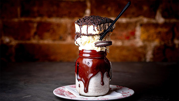 A decadent dessert featuring a creamy milkshake in a jar, topped with dripping chocolate sauce, a large slice of cake, whipped cream, a cookie, and a chocolate straw, placed on a white plate with red drizzle, against a rustic brick wall background.