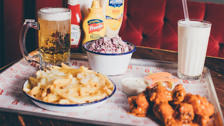 A tray with a variety of food items including a bowl of fries, a plate of saucy chicken wings, and a bowl of coleslaw. Drinks include a glass mug of beer and a glass of milkshake with a straw. Condiments like ketchup and mustard are placed in the background.