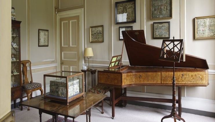 A cozy room featuring an angled wooden spinet against the wall, framed artworks hanging above it. A display case with intricate embroidery sits atop a wooden table in the foreground, next to a music stand. A chair with a floral cushion is placed near a corner cabinet.