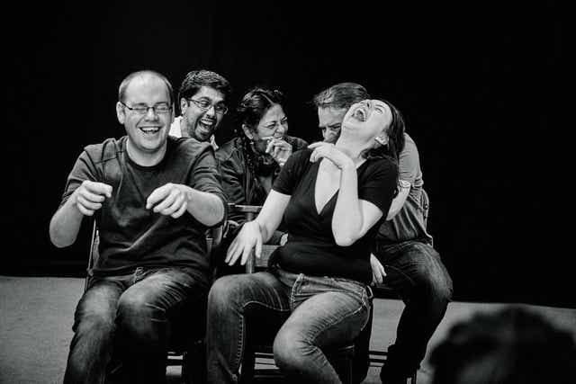 A black and white photo of five people sitting closely together and laughing heartily. The mood appears joyful and lighthearted, and their varying expressions and gestures suggest they might be reacting to something humorous or entertaining.