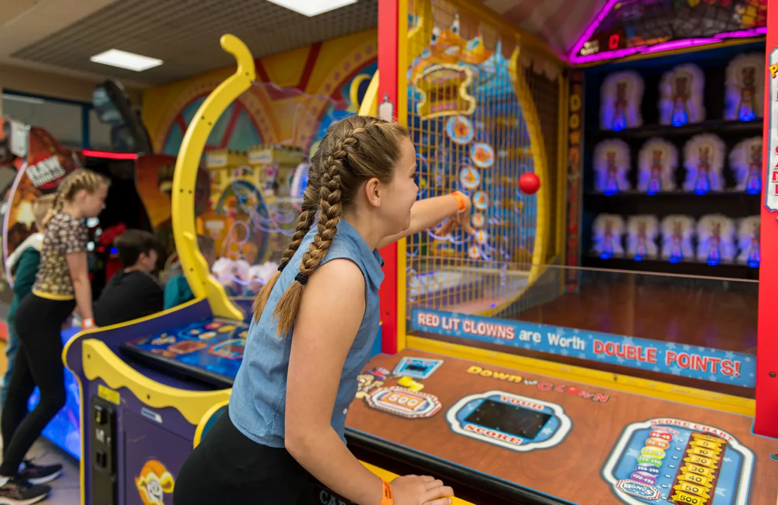 A young girl with braided hair throws a red ball at a target in an arcade game. She is standing in front of a colorful game machine with various lights and displays. Other arcade machines and players are visible in the background.