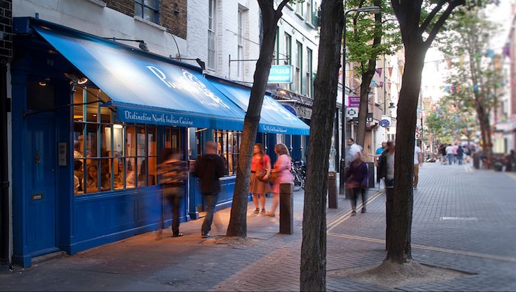 A lively street scene features people walking past a blue storefront with an awning. The setting is a narrow, tree-lined pedestrian street with a relaxed atmosphere, where the soft evening light enhances the vibrant yet calm environment.