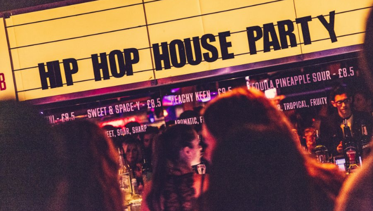 A vibrant, crowded bar with a prominent yellow sign above that reads HIP HOP HOUSE PARTY. People are mingling and enjoying drinks in the foreground, while bartenders work behind the counter. Cocktail names and prices are partially visible on the sign.