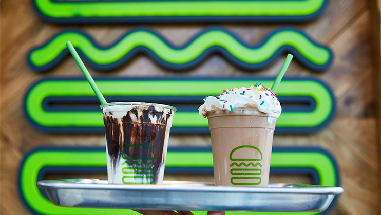 Two milkshakes on a metal tray. The one on the left has chocolate drizzle, and the one on the right has whipped cream with rainbow sprinkles. Both shakes have green straws and are in cups with a logo. A wall with a green wavy design is in the background.