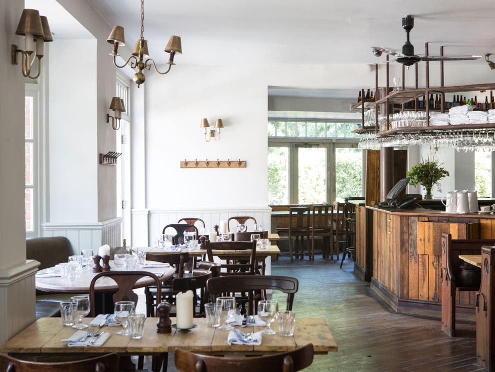 A cozy restaurant interior featuring wooden tables set with glassware and white napkins. The space has a rustic charm with wooden floors, a wooden bar, and soft lighting from wall-mounted lamps and chandeliers. Large windows let in natural light.