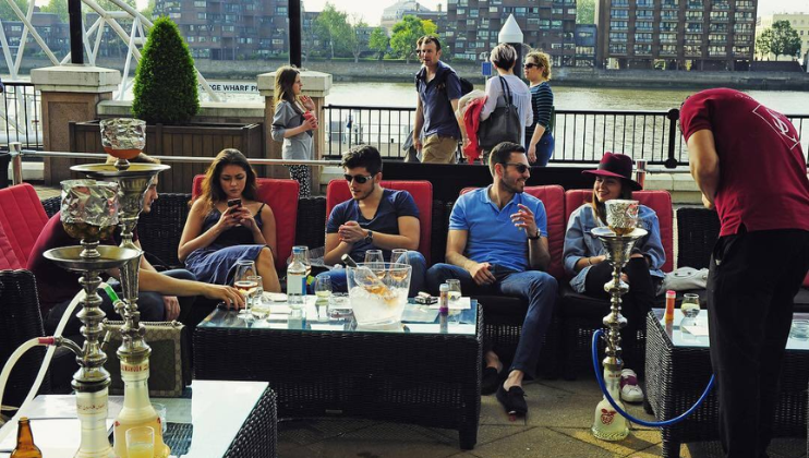 A group of people sit on outdoor couches next to a river, smoking from hookahs and chatting. Three people on the left focus on their phones while two others engage in conversation. A server in a red shirt attends to the hookahs. Other people walk in the background.