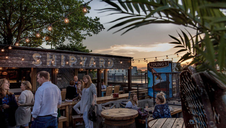 Outdoor bar area at sunset. Cozy wooden seating with tables and string lights create a warm ambiance. Large sign reads SHIPYARD. People are casually gathered, socializing and enjoying the evening. Green foliage and a YOUNG'S sign are visible.