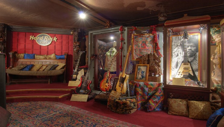 A cozy corner of Hard Rock Cafe adorned with musical memorabilia. Various guitars and framed photos are on display, along with vintage decor and colorful fabric drapes. A seated area with plush cushions is visible to the left.