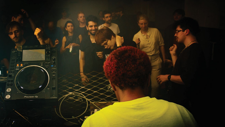 A DJ with bright red hair is performing in a dimly lit nightclub. Behind and around the DJ, a diverse group of people is dancing, some holding drinks and others talking or cheering. DJ equipment is visible in the foreground, with a crowd enjoying the music in the background.