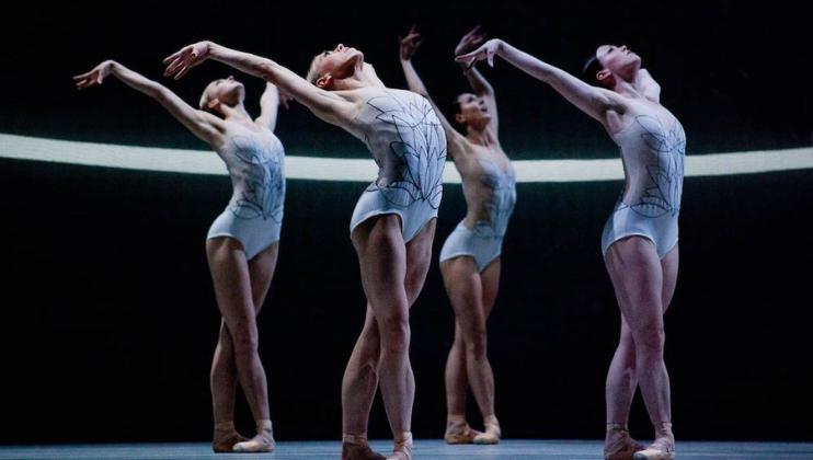 Against a black background, four female ballet dancers in white leotards, arranged in a diamond formation, stretch their arms upwards and back