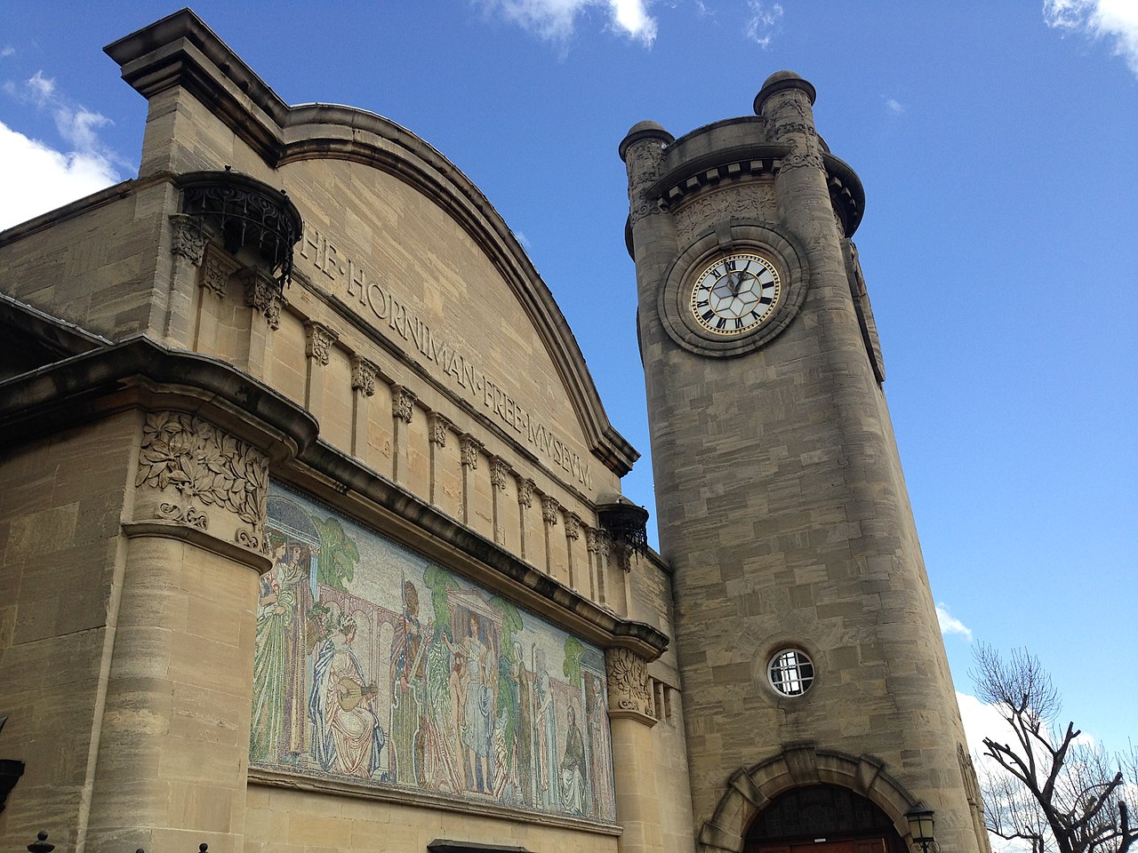 A historic stone building with a round clock tower stands against a partly cloudy blue sky. The facade features an intricate mosaic depicting people in classical attire. The building displays an inscription in a classical style above the mosaic.