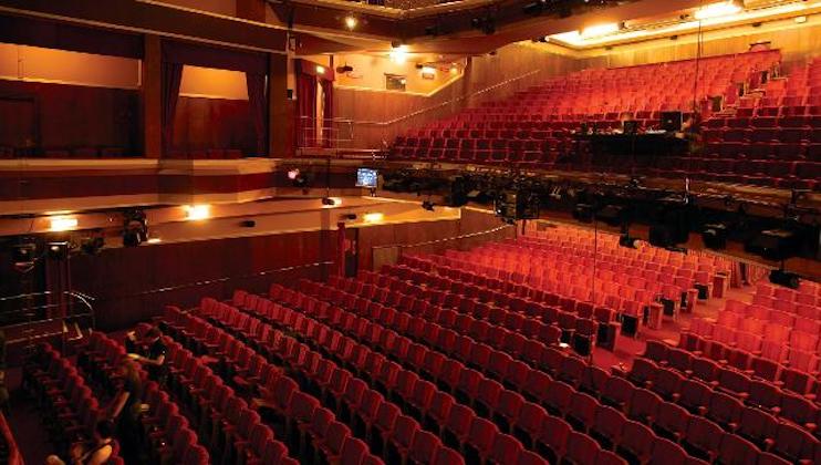 A large, empty theater with rows of red seats facing a stage. Warm lighting from chandeliers and wall fixtures illuminates the space. The stage is equipped with various lighting rigs and curtains, creating a cozy and inviting atmosphere.