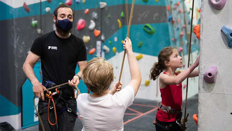 A bearded man wearing a face mask and a black shirt assists two children climbing an indoor rock wall. The boy in a white shirt is holding a rope, while the girl in a red shirt begins her climb. Colorful climbing holds cover the wall in the background.