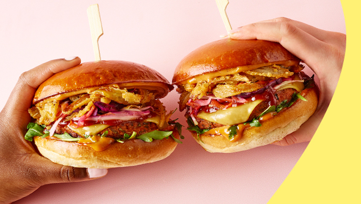 Two hands holding large gourmet burgers with glossy buns, filled with melted cheese, crispy fried onions, lettuce, red onions, and vibrant shredded vegetables, against a pink background with a curved yellow accent.