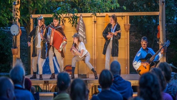 A group of five musicians perform on an outdoor wooden stage under trees. They play various instruments, including an accordion, guitar, and ukulele. The audience watches attentively from their seats. The stage is lit warmly, creating a cozy atmosphere.