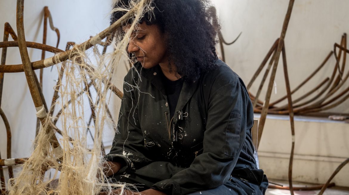 A person with curly hair sits in a workshop, wearing a dark jumpsuit. They are weaving or handling fibers among rusted metal structures. Sunlight filters into the space, illuminating their focused expression and highlighting the textures around them.