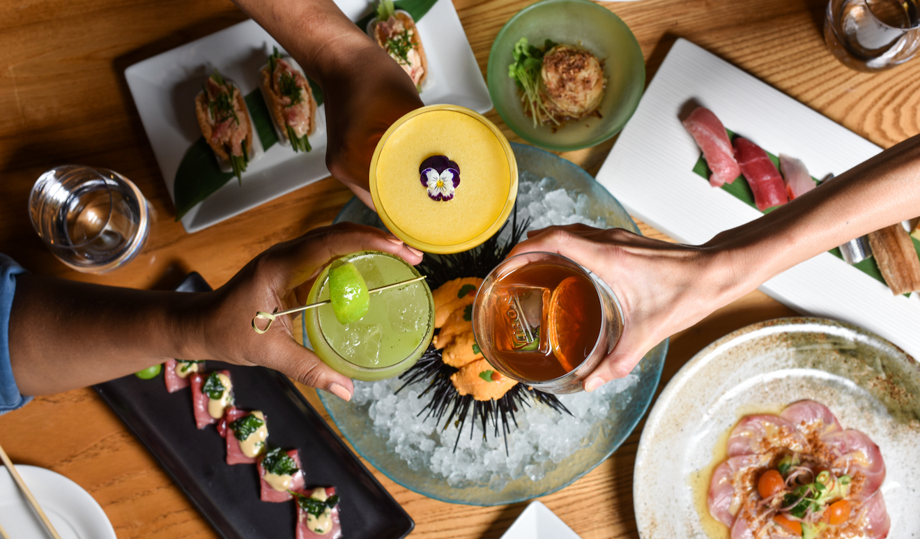 Four hands holding colorful cocktails over a table filled with plates of sushi and other gourmet dishes. The table is set on a wooden surface, showcasing a vibrant and diverse spread of food, including sashimi, nigiri, and a dish served on ice.