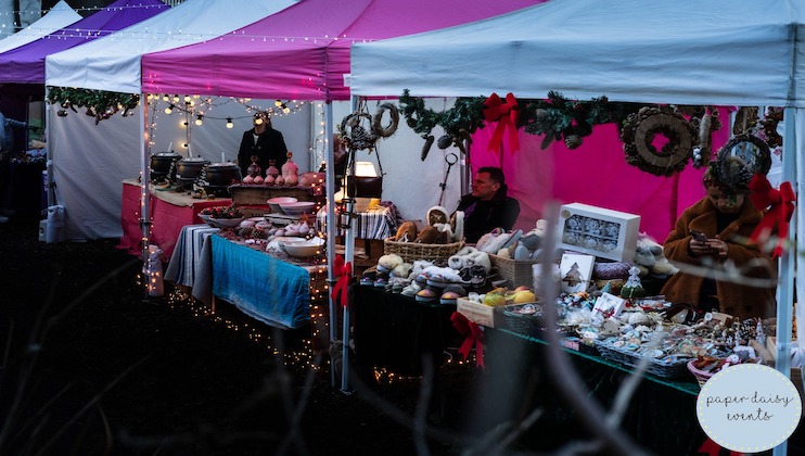 A festive market scene with brightly colored stalls displaying various items. One stall, adorned with wreaths and Christmas lights, features wool products and decorations. Two vendors are present, one standing and one seated, tending to their displays.