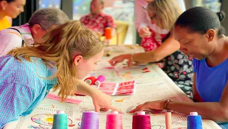 A diverse group of people, including adults and children, are gathered around a table engaged in a crafting activity. Various colorful spools of thread, markers, and paper are spread out. A young girl and an adult woman intently focus on a shared project.