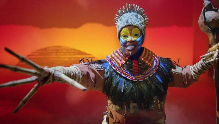 A performer from The Lion King musical, wearing elaborate face paint, headdress, and vibrant tribal costume, extends their arm dramatically against a backdrop of a stylized sunset over an African landscape.