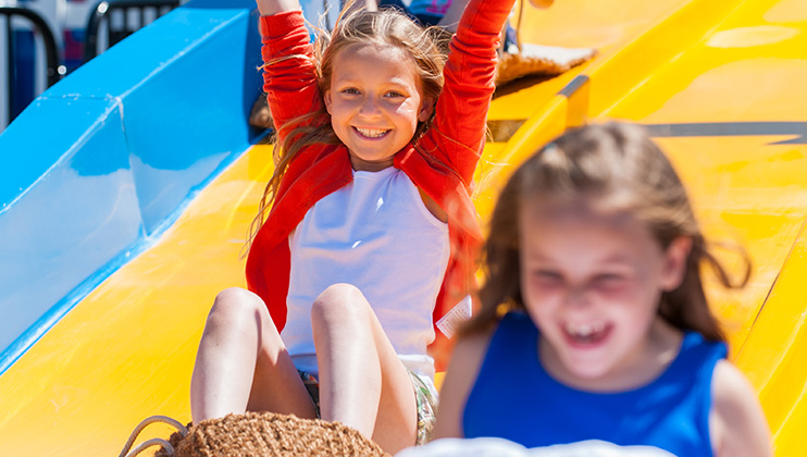 Two children are enjoying a ride on a colorful slide. The child in front, wearing a red and white top, is smiling with arms raised, while the child in blue in the foreground is also smiling. They appear to be sliding down on mats on a sunny day.
