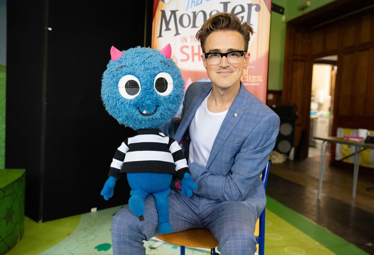 A man dressed in a light blue plaid suit and white t-shirt sits on a chair holding a blue puppet with a round head, horns, and big eyes. The puppet is wearing a black and white striped shirt. They are in front of a colorful poster with the word Monster.