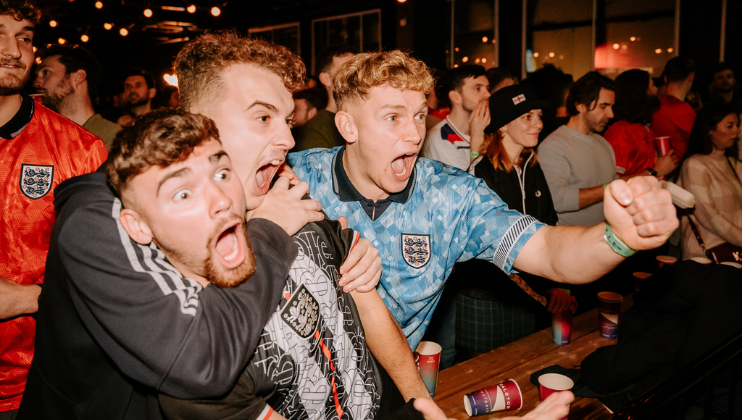 A group of excited people, mostly dressed in football jerseys, are passionately cheering and celebrating in a lively bar. In the foreground, three men are in animated expressions, with one in a blue jersey pointing forward. The bar is dimly lit with warm lights.