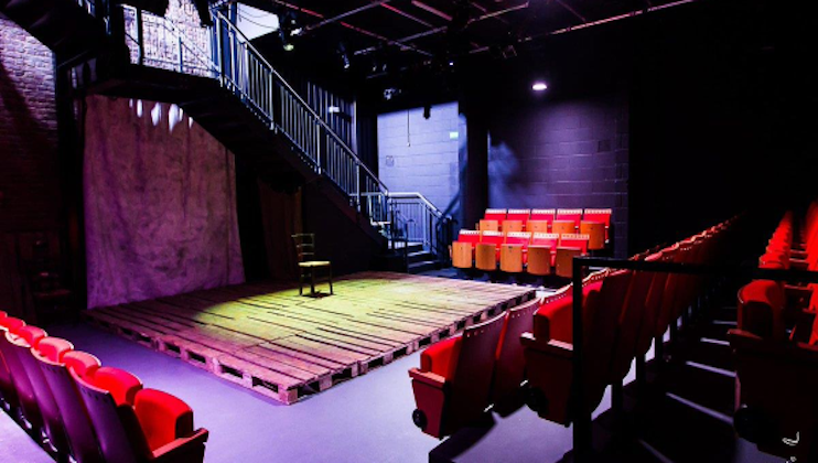 A small theater with a wooden pallet stage illuminated by spotlight. The space features tiered red seating on the right and at the back, and a staircase leading to a raised area on the left. The ambiance is dark with dramatic lighting.