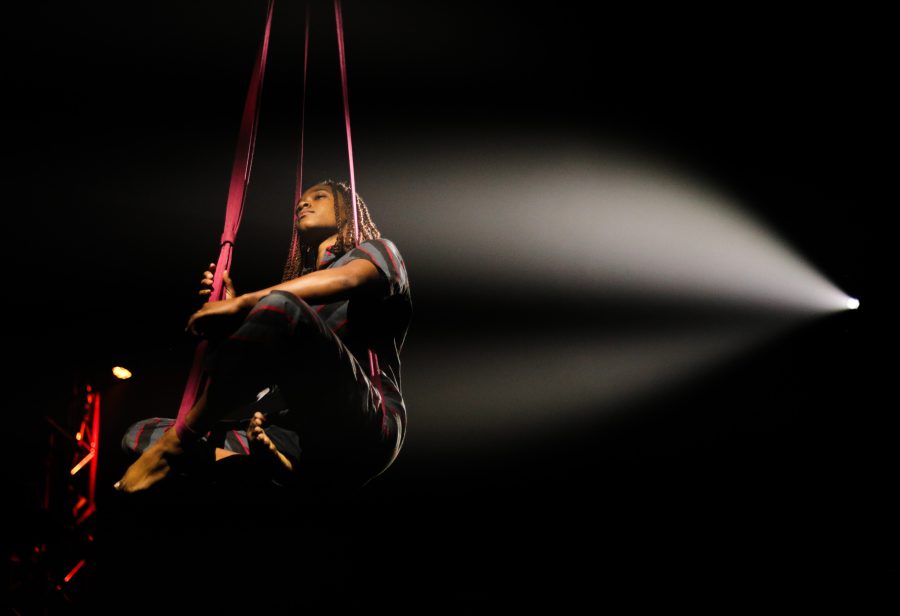A person performs an aerial acrobatics routine suspended from two long, pink ribbons in a dimly lit scene. They are illuminated by a spotlight from the right, highlighting their focused expression and skillful balance. The background is mostly dark with hints of stage lights.