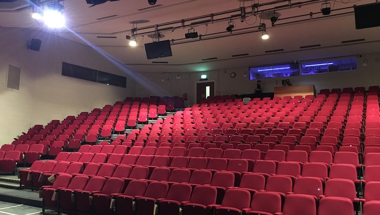 A large auditorium with tiered seating featuring rows of empty red chairs. The room is dimly lit with a few overhead lights, and there is a control room with a glass window visible at the back. A few television screens hang from the ceiling.