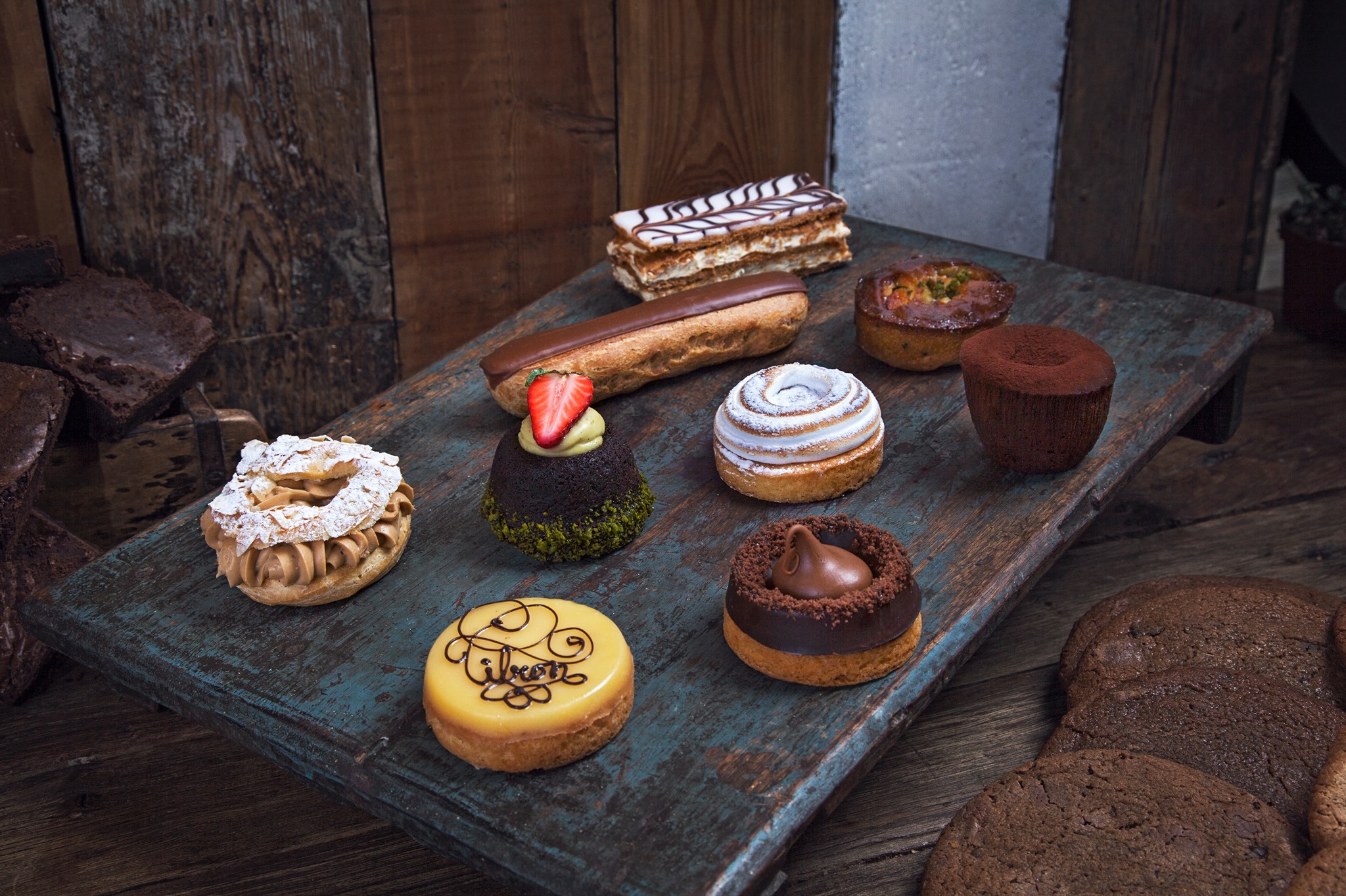 A rustic wooden board displays an assortment of pastries, including éclairs, tarts, cakes, and cookies. Each pastry is uniquely decorated, with some featuring fruits, cream, and chocolate embellishments, creating a visually appealing and diverse dessert spread.