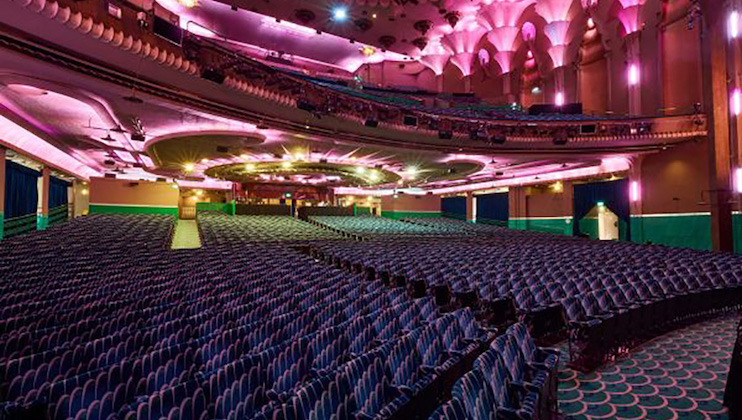 A lavish, empty theater auditorium with rows of blue seats and an ornate ceiling featuring pink and purple lighting. The stage is not visible in this angle, but the upper balcony section and detailed architectural design suggest an elegant and expansive space.