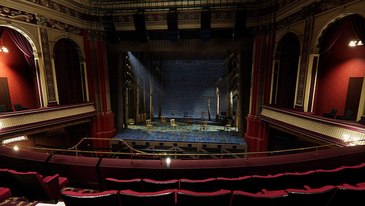 A grand, historic theater with ornate details and red velvet seating. The stage is set with rustic wooden chairs and minimal props, lit dramatically by spotlights. Golden balconies on either side of the theater add to the opulent atmosphere.