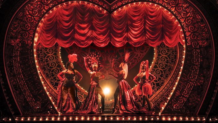 Four performers in elaborate costumes, including feathers and sequins, stand on a heart-shaped stage with ornate decorations. The stage is adorned with red drapes and warm lighting, creating a vibrant and lively atmosphere reminiscent of a theatrical or cabaret performance.