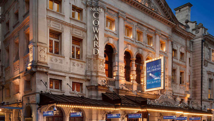 The image shows the front facade of the Noël Coward Theatre in London. The building exterior features ornate architectural details with large arched windows and decorative stonework. Signs reading Delfont Mackintosh Theatres and Noël Coward are prominently displayed.