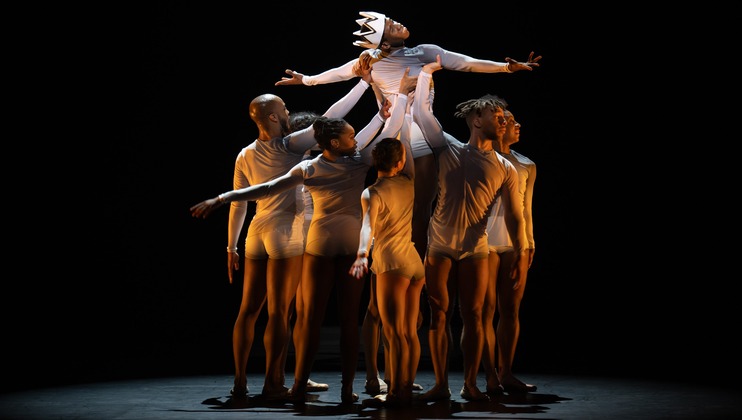 A group of dancers wearing white outfits perform a contemporary dance on stage. One dancer, wearing a crown, is lifted above the others, extending their arms outward. The scene is illuminated against a dark background, creating a dramatic effect.