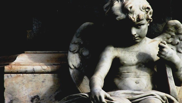 A close-up of a marble statue of a cherub with wings. The cherub is leaning forward, holding an object and appears deep in thought. The lighting casts shadows, creating a dramatic and somber mood. The background is dark, highlighting the details of the statue.